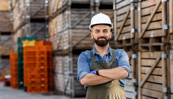 Inspecting products in warehouse, management, distribution and logistics. Happy confident young bearded man in uniform and hard hat with crossed arms, on many wooden boxes with goods background